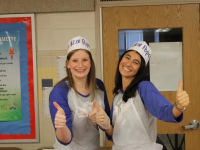 Two teachers manning a food table with thumbs up