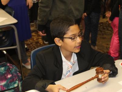 A boy in suit jacket sits at a desk holding a gavel.